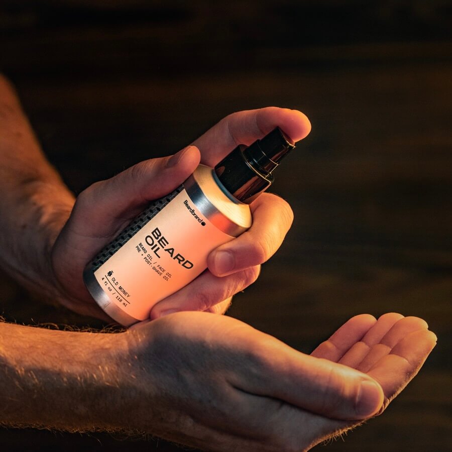 A person holding in their hand a bottle of Beardbrand Old Money Beard Oil and showing the front label. They are about to pump the product into their other hand.